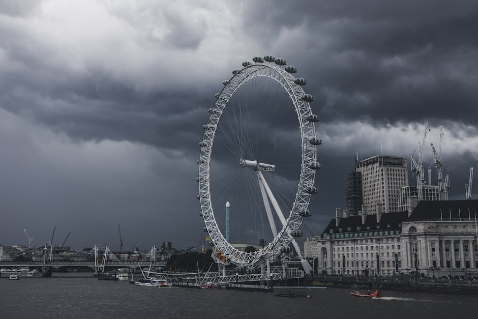 A large Ferris wheel situated along the riverbank under a dark, cloudy sky. The wheel has enclosed passenger cabins evenly spaced around its circumference, with some cabins visible on the upper and lo