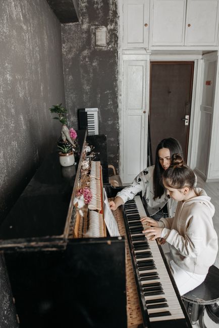 Inside a residential room with light-colored paneled walls and a parquet wooden floor, there is an upright wooden piano positioned against the wall, with an open sheet music book resting on its music