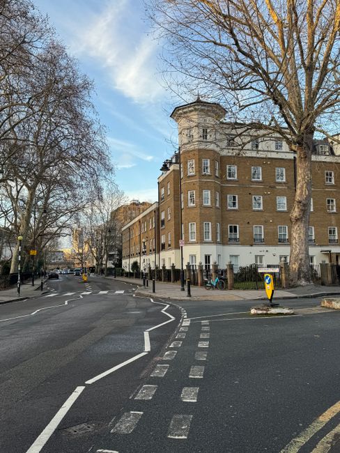 A wide street view in Lambeth featuring a large beige and brown residential building with multiple windows and a small turret on the corner. Several leafless trees line the pavement, casting shadows o