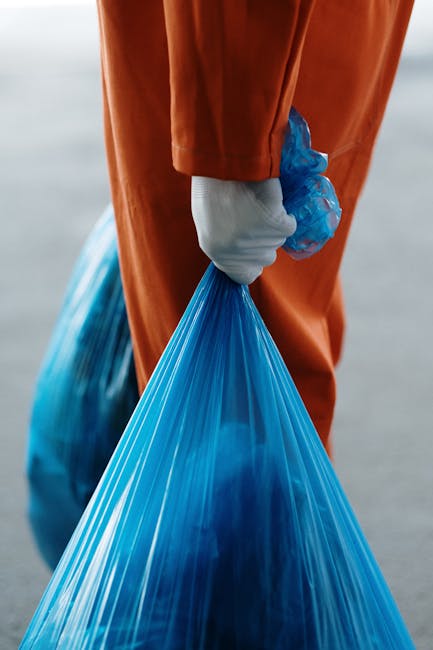 A sanitation worker wearing a high-visibility yellow and red uniform is emptying a large red waste collection truck parked on the side of a street. The truck's rear hopper is open, revealing stored wa